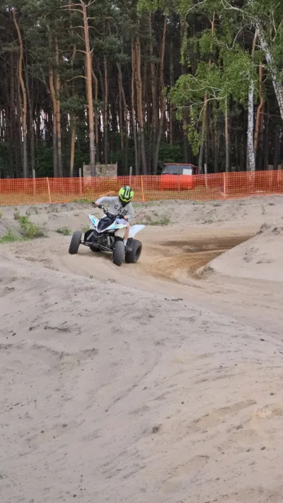 Quad na piaszczystym torze, w tle pomarańczowa siatka i sosny